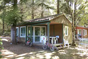 Campinghaus im Wald mit überdachter Terrasse, weißen Stühlen und Tisch sowie Fahrrädern vor der Tür.