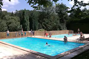 Schwimmbecken im Campingbereich mit Holzgeländer, Menschen im Wasser und auf der Liegefläche, umgeben von Bäumen.