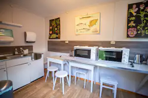 Küchenzeile im Campingbereich mit Spüle, Wasserkocher, Mikrowelle und Backofen, dazu zwei weiße Hocker und Wandbilder.