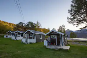 Reihe von Mobilheimen auf einem grünen Campingplatz, mit Holzveranda, Stufen und Weg entlang der Straße, Bäume im Hintergrund.