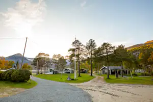 Campingplatz mit Kiesweg, mehreren Mobilheimen und Wohnwagen, Bäumen und einem Auto am Rand, im Hintergrund Hügel.