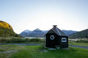 Holzhütte auf einem Campingplatz im Grünen, mit Rasenfläche, Büschen und Blick auf Berge im Hintergrund.