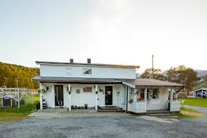 Campinghaus mit Veranda, Holzgeländer und Eingangstür, davor Kiesweg und Rasen, im Hintergrund weitere Gebäude und Bäume.