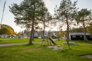 Spielplatz mit Schaukel und Rutsche auf einer grünen Wiese, umgeben von Bäumen und Camping-Unterkünften.