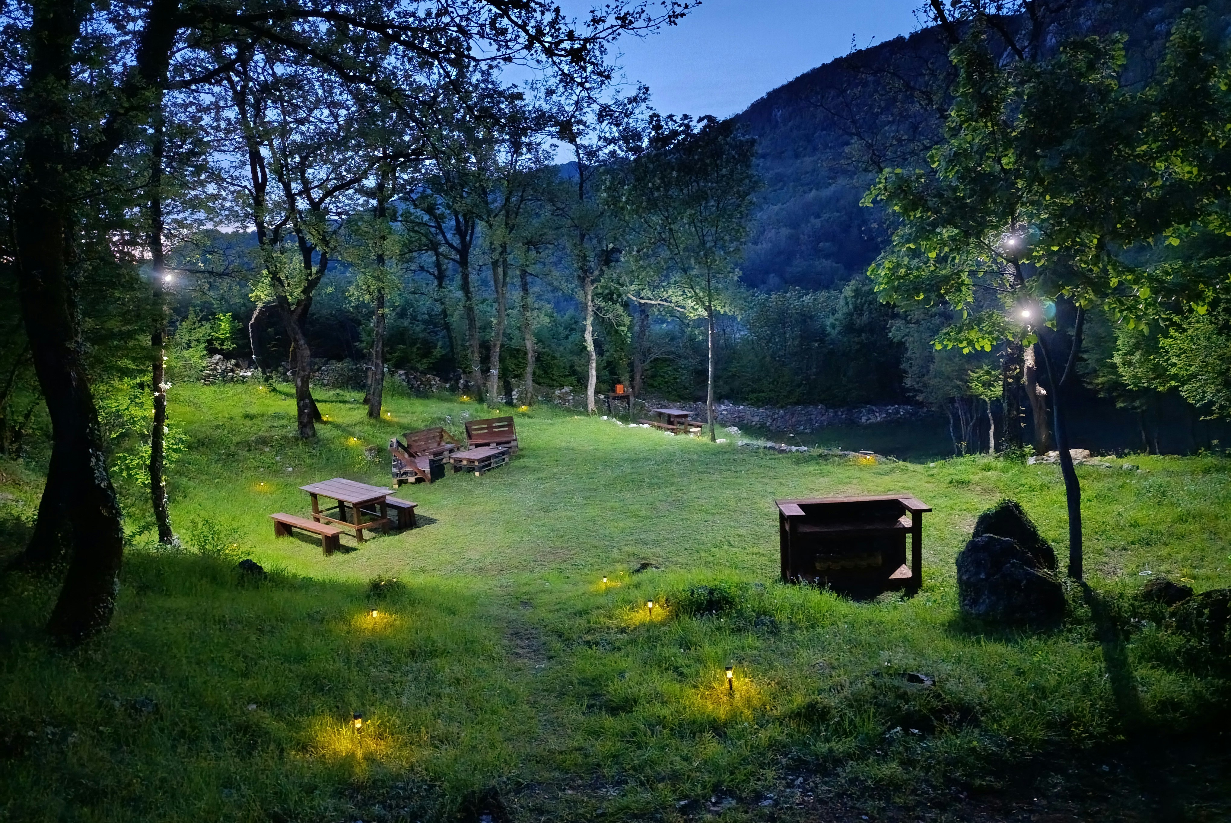Campingplatz im Abendlicht mit mehreren Holz-Tischen, Feuerstellen und einem kleinen Schuppen am grünen Hang zwischen Bäumen.
