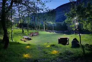 Campingplatz im Abendlicht mit mehreren Holz-Tischen, Feuerstellen und einem kleinen Schuppen am grünen Hang zwischen Bäumen.
