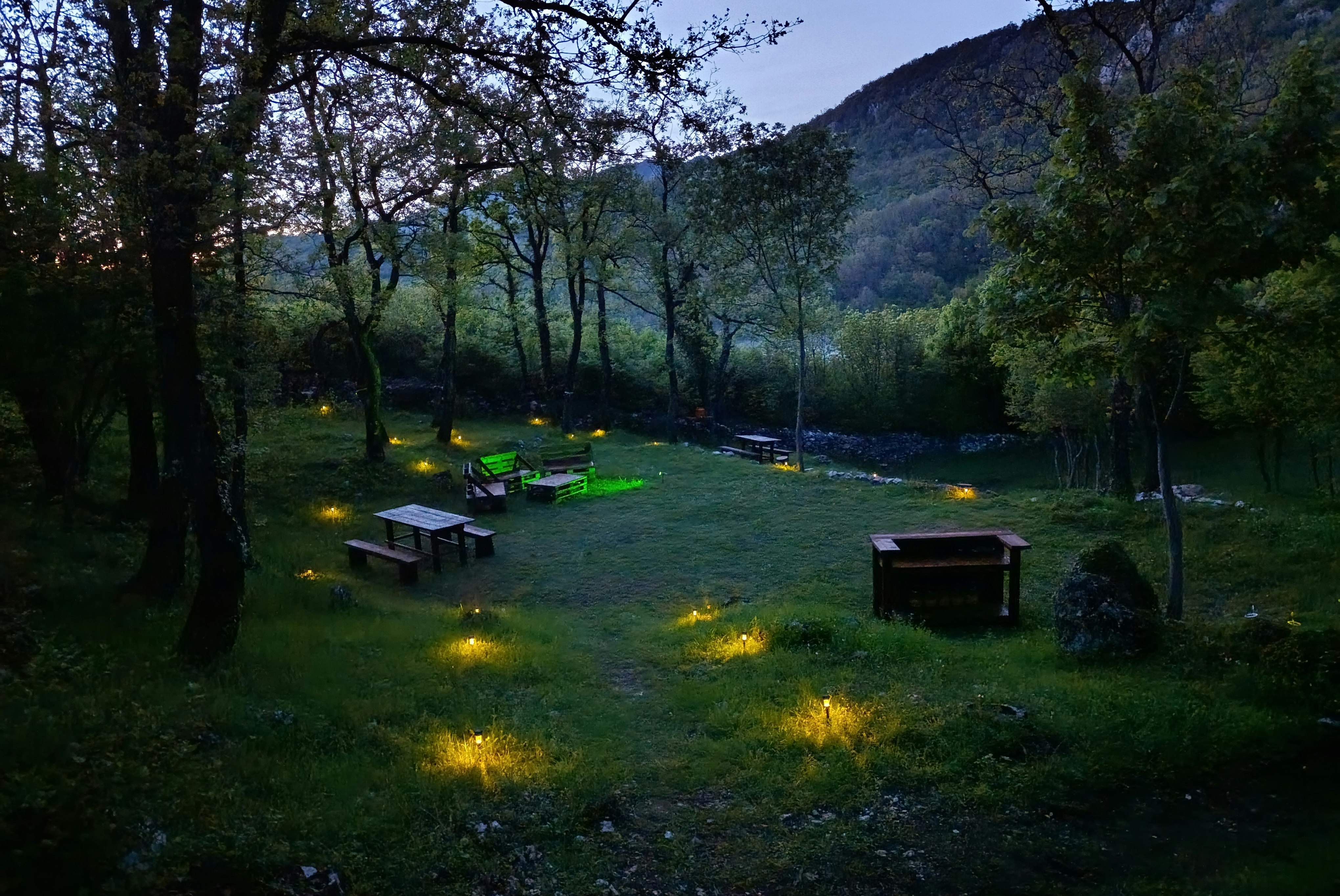 Campingplatz im Grünen mit mehreren Grillstellen und Picknicktischen, umgeben von Bäumen und einem Hang im Hintergrund.