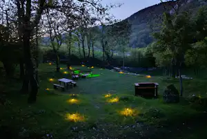 Campingplatz im Grünen mit mehreren Grillstellen und Picknicktischen, umgeben von Bäumen und einem Hang im Hintergrund.