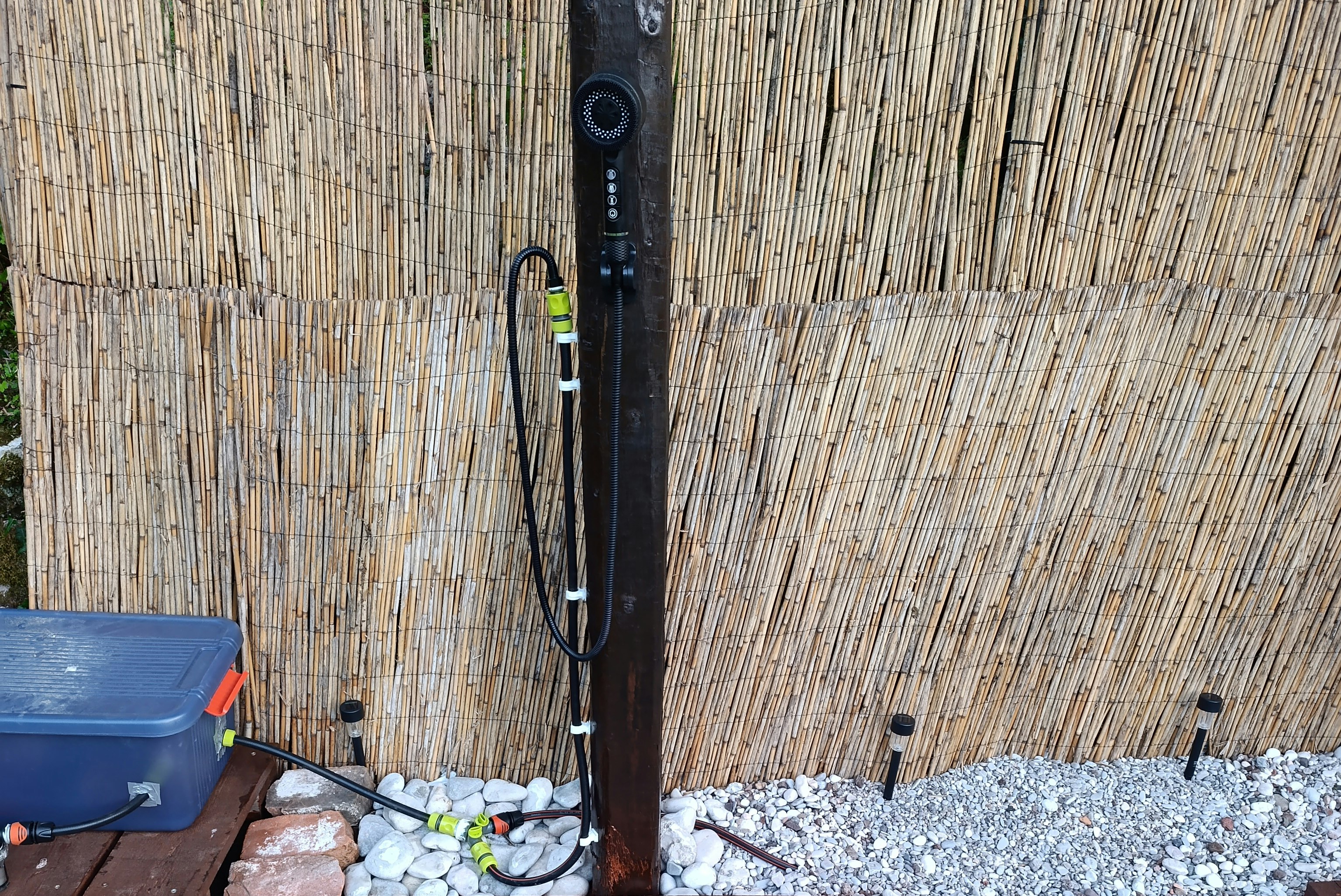 Außendusche an einer Bambuswand mit Bodenwanne aus Metall und Kiesfläche, daneben Pflastersteine.