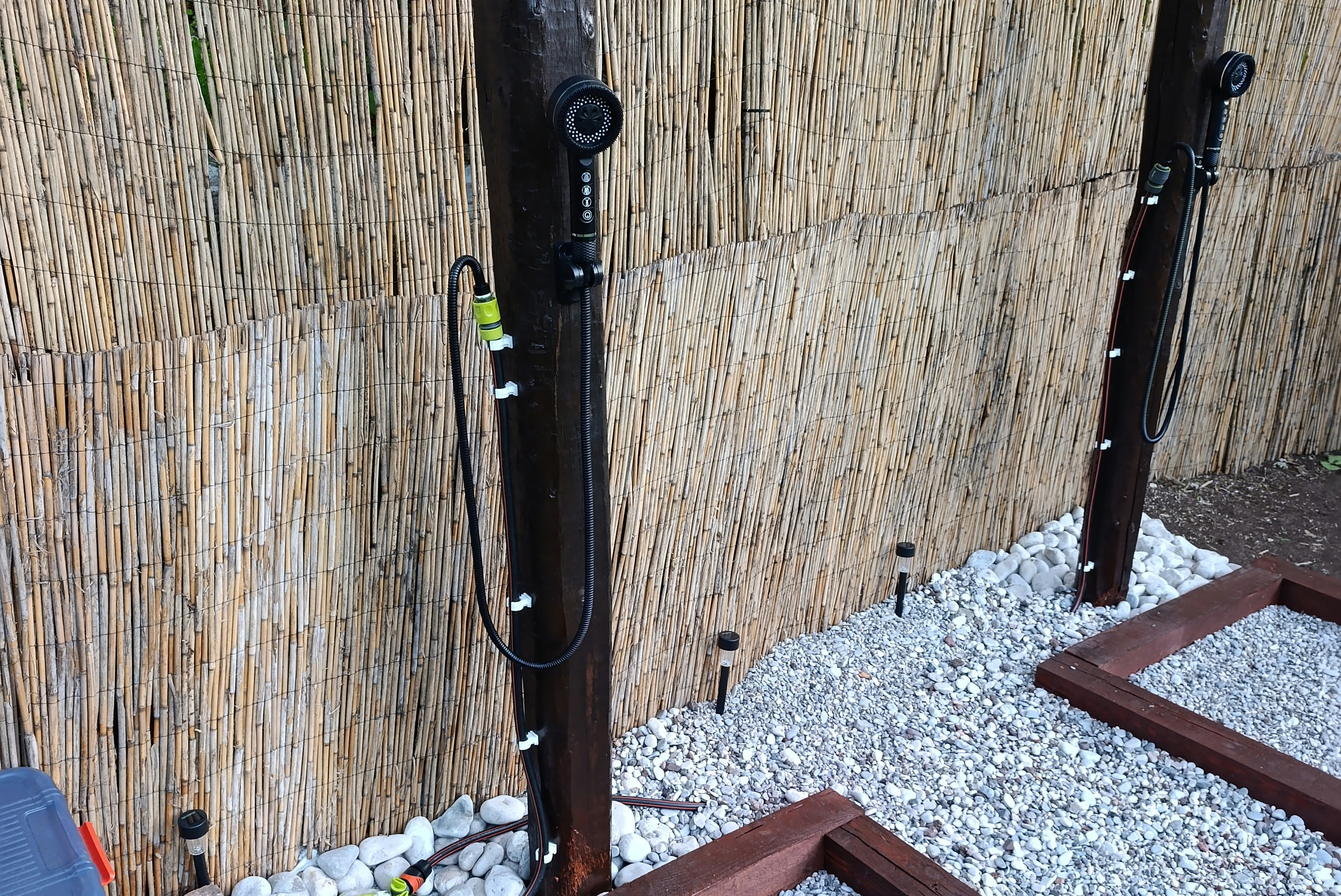 Außendusche am Campingplatz mit Bambuszaun, Kiesboden und Holzrahmen, daneben Steine und ein kleiner Wasserhahn.