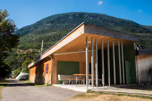 Campinggebäude mit überdachter Terrasse, Holzbalken und Tisch, davor Parkplatz und Auto, im Hintergrund bewaldeter Berg.