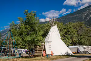 Zelt auf einem Campingplatz mit Holzleiter, daneben Wohnwagen und Auto, im Hintergrund Bäume und Berge.