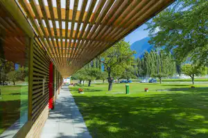 Holzveranda am Campingplatz mit Schattenstreifen, gepflastertem Weg, Rasen und Bäumen; im Hintergrund Berge.