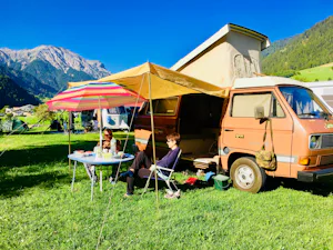 Camping Muglin - Camper sitzen vor dem Wohnmobil im Schatten der Markise