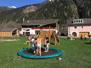 Trampolin auf dem Campingplatz mit Kindern vor Holzterrasse und Spielbereich, im Hintergrund Berge und Waldhang.