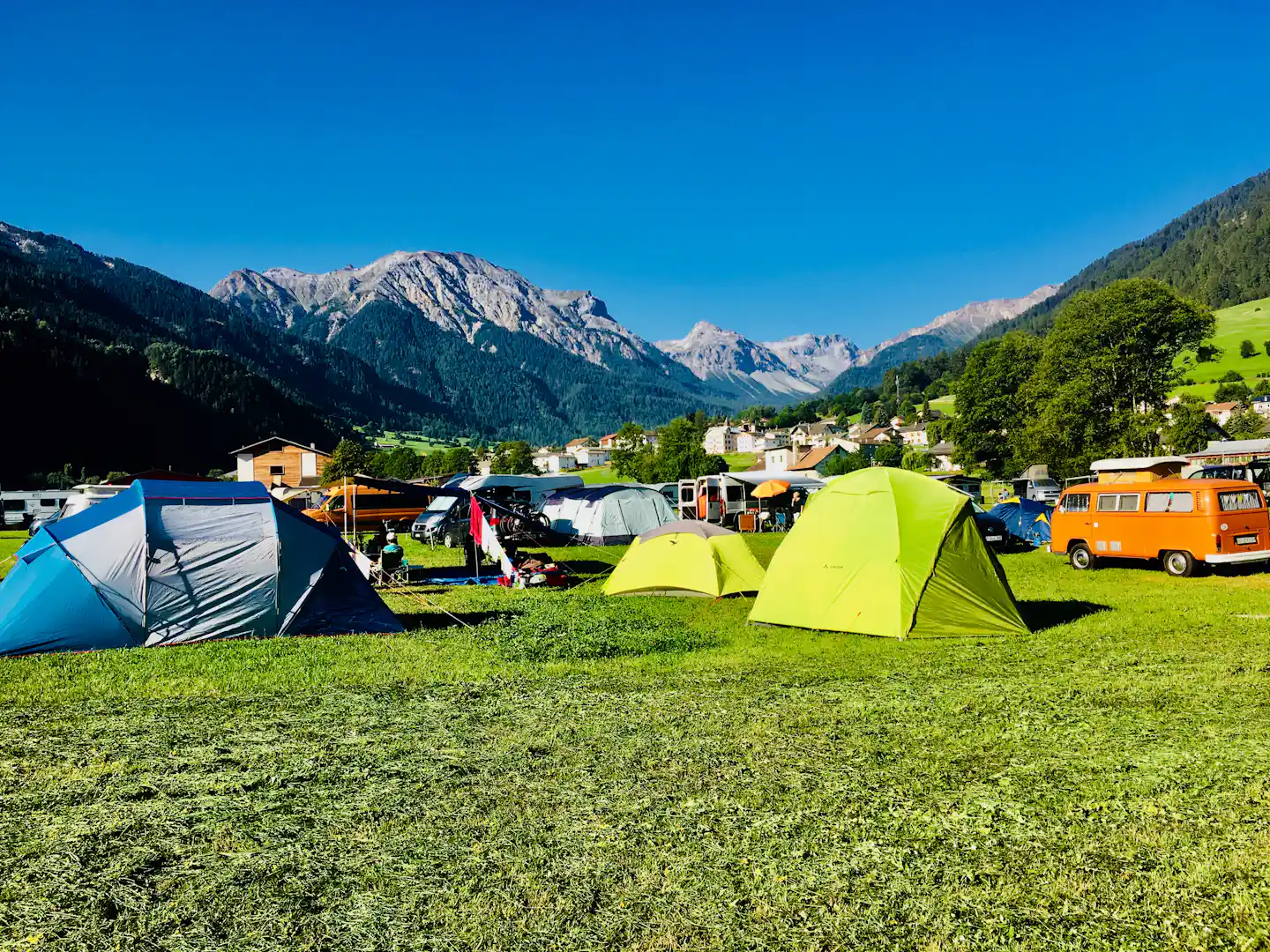 Campingzeltplatz mit bunten Zelten, Autos und Wohnmobilen, im Hintergrund Alpen und ein Dorf am Hang.