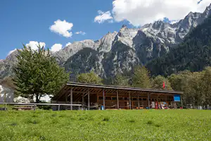 Campinggebäude mit überdachter Terrasse am Hang, umgeben von Wiesen und Bäumen, im Hintergrund hohe Alpenfelsen.