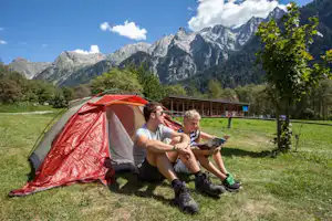 Campingzelt in Rot auf einer Wiese, daneben sitzen zwei Personen am Tisch vor einem Holzgebäude mit Bergblick.