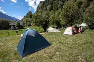 Campingzelte auf einem Grasplatz, im Hintergrund Bäume, Berge und ein kleiner Schuppen am Rand des Campingplatzes.