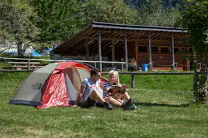Campingzelt auf grünem Rasen neben einem Holzunterstand, zwei Personen sitzen davor und schauen auf ein Tablet.