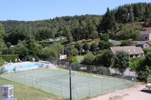 Tennisplatz und Pool auf einem Campingplatz, umgeben von grünen Hügeln, Bäumen und mehreren Gebäuden im Hintergrund.