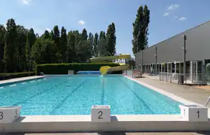 Schwimmbecken am Campingplatz mit Startblöcken, daneben ein Gebäude mit Geländer und Bäumen im Hintergrund.