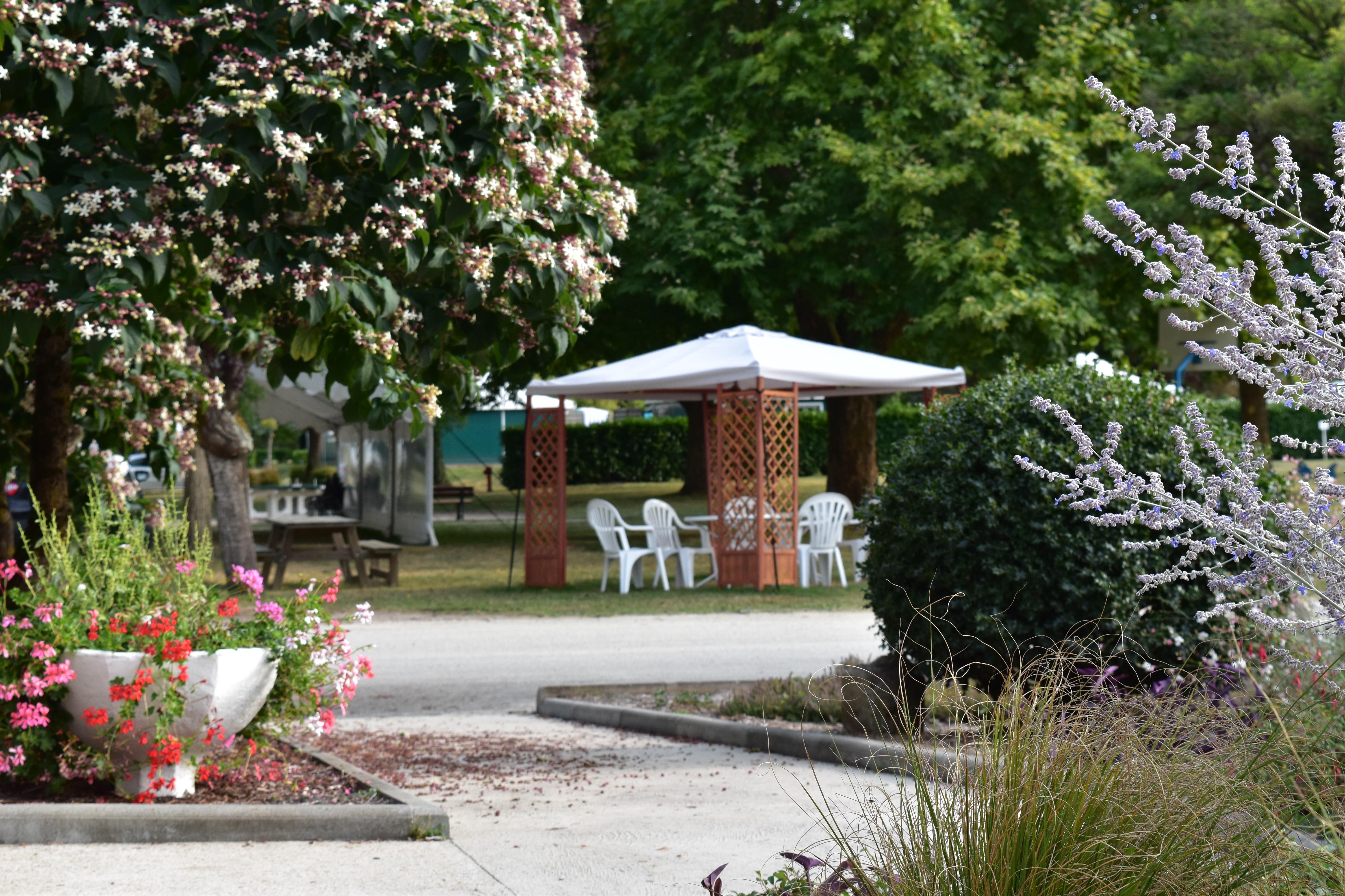 Blumenbeete und Kiesweg im Campinggarten mit Pavillon und Sitzgruppe unter weißem Sonnenschirm.