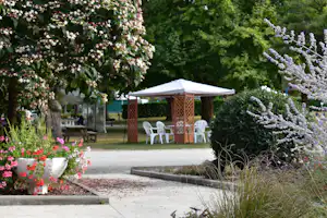 Blumenbeete und Kiesweg im Campinggarten mit Pavillon und Sitzgruppe unter weißem Sonnenschirm.