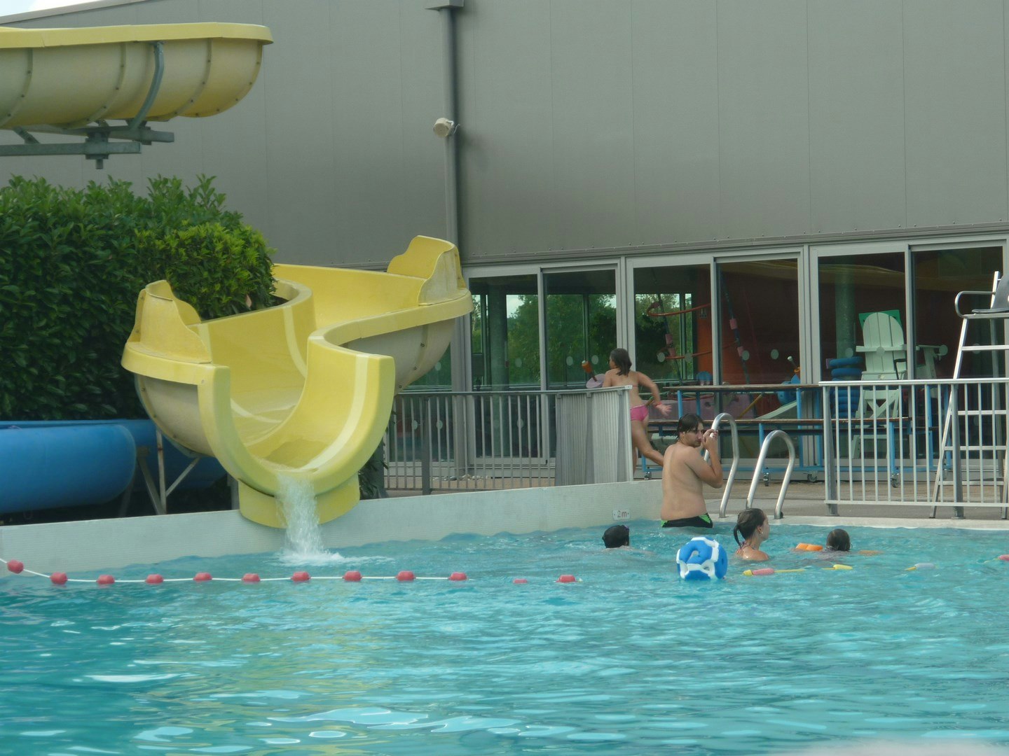 Familienpool mit gelber Rutsche, blauen Spielzeugen im Wasser und Zaun vor einem modernen Campinggebäude.