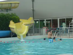 Familienpool mit gelber Rutsche, blauen Spielzeugen im Wasser und Zaun vor einem modernen Campinggebäude.