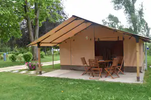 Großes Campingzelt mit Holzveranda und Sitzgruppe auf dem Stellplatz, umgeben von Rasen und Bäumen.