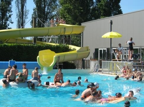 Kinder und Erwachsene im Camping-Schwimmbad mit gelber Rutsche vor einem weißen Gebäude und Metalltreppe.