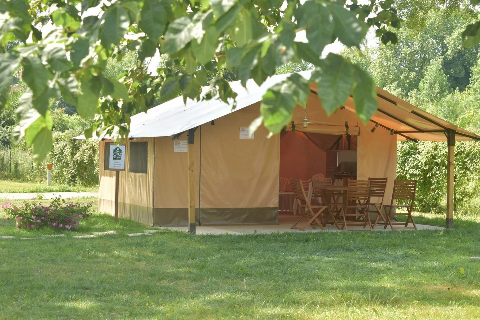 Zelt mit Holzstühlen und Tisch auf einem grünen Rasen, umgeben von Bäumen und Hecken im Campingbereich.