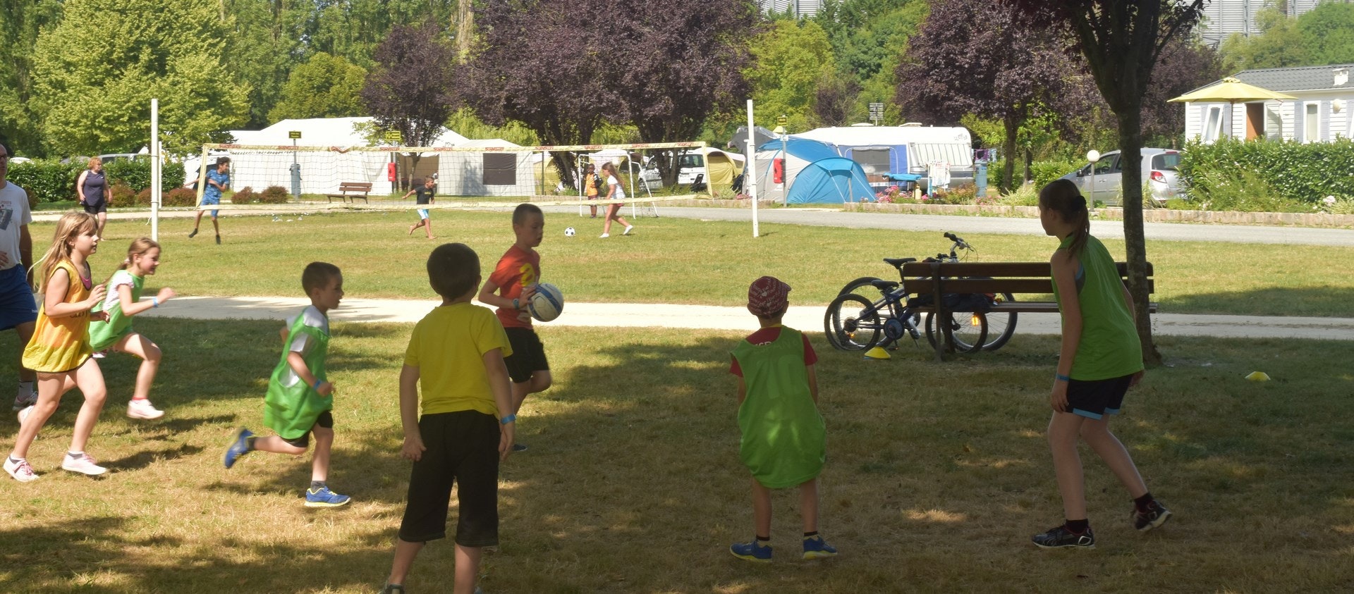 Kinder spielen auf einer Wiese im Campingpark, im Hintergrund Zelte und Wohnwagen sowie ein Spielplatz mit Schaukeln.