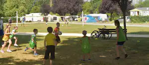 Kinder spielen auf einer Wiese im Campingpark, im Hintergrund Zelte und Wohnwagen sowie ein Spielplatz mit Schaukeln.