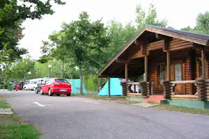 Holzblockhütte mit Veranda und Sitzbereich, daneben Zelt und geparkte Autos auf dem Campingplatz im Grünen.
