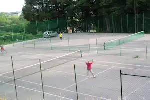 Tennisplatz auf dem Campinggelände mit grünen Zäunen, Bäumen im Hintergrund und spielenden Kindern mit Tennisbällen.