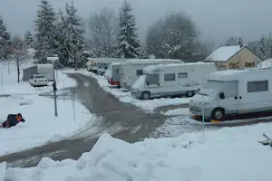 Wohnmobile auf einem Campingplatz im Schnee, schmale Straße zwischen Stellplätzen, Bäume und Gebäude im Hintergrund.