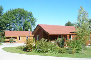 Holzhaus mit rotem Ziegeldach und überdachter Terrasse, Blumenbeete und Bäume im Hintergrund.