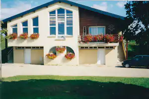 Ferienhaus mit Terrasse, Blumenampeln und großen Fenstern, davor Rasen und ein geparktes Auto am Rand.