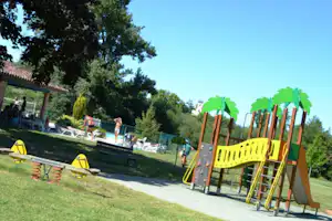 Bunter Spielplatz am Campingplatz im Grünen mit Rutsche, Schaukeln und Rasenfläche, im Hintergrund Liegen und Bäume.
