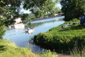 Blick auf den See mit Grasufer, Bäumen und einem kleinen Boot; am Rand liegen Liegestühle und ein Auto.