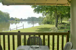 Blick vom Terrassenplatz auf einen ruhigen See, Rasen und Bäume; Tisch und Stühle unter einem grünen Sonnendach.