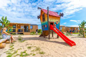 Spielplatz am Campingplatz mit roter Rutsche, Holzspielhaus und Sandfläche vor mehreren Holzhäusern.