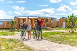 Zwei Personen schieben Fahrräder auf einem Kiesweg vor Holzbungalows, mit Rasenfläche und Bäumen im Hintergrund.