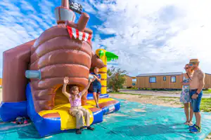 Aufblasbares Hüpfburg-Spielgerät mit Rutsche und Poolbereich, daneben ein Campingplatzgebäude und Personen am Wasser.