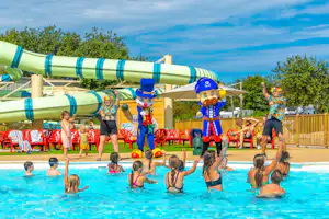 Kinder und Erwachsene im Pool vor einer großen Wasserrutsche, im Hintergrund Sonnenschirme und Spielbereich am Campingplatz.