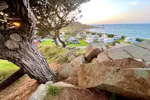 Campingplatz am Meer: Stellplätze mit Wohnwagen und Bäumen am Hang, im Hintergrund die Küste und das Wasser.