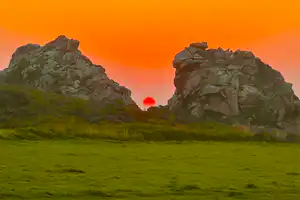 Grüne Wiese vor zwei Felsbergen, die Sonne steht tief am orangefarbenen Himmel beim Camping-Landschaftsblick.