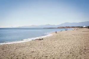 Sandstrand am Meer mit Blick auf Berge, Badegäste und ein weißes Zelt am Ufer.